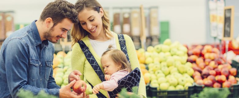A couple and their young toddler stroll through the produce aisle in the supermarket and show their child an apple.