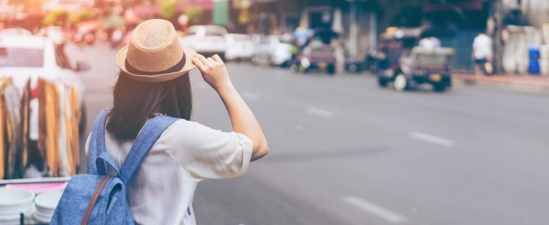 A woman wearing a fedora and a backpack, about to cross the street.