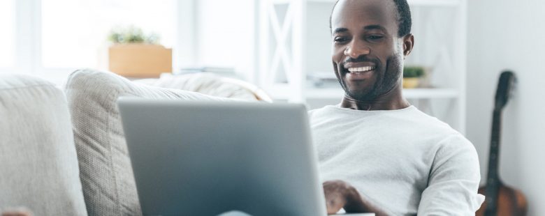 A man sits on a couch smiling at his laptop.