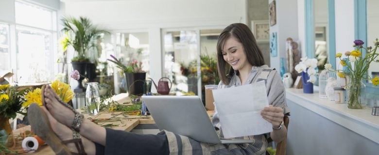 A woman with her feet up, looking at her laptop and some papers, smiling because she's mastered budgeting basics.