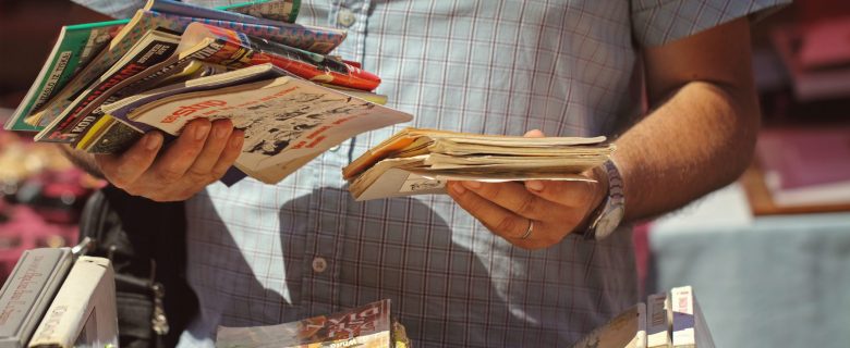 A man stands in front of a table stacked with books holding comic books in both of his hands. 