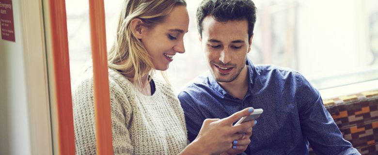 A man and a woman sitting together on a couch, looking at something on a smart phone.