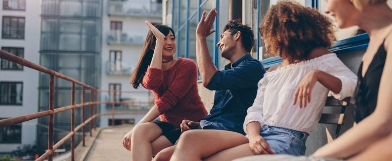 Friends enjoying a summer day on the patio, surrounded by other buildings. Two people laugh as the other two high five one another.