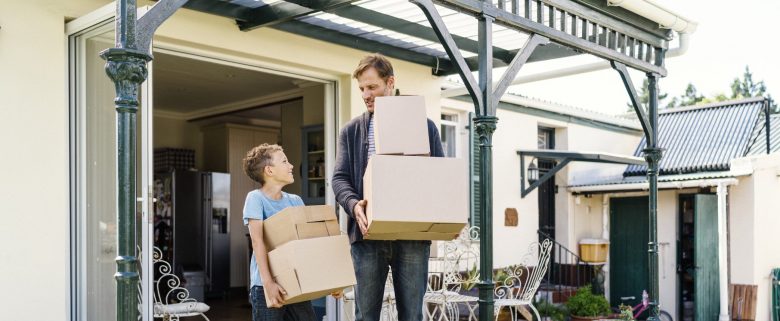 Man and child smiling at one another as they walk outside carrying boxes from a house.