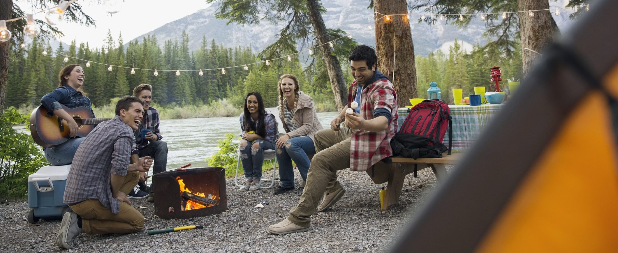 A group of friends, camping by a river, sitting around a campfire.