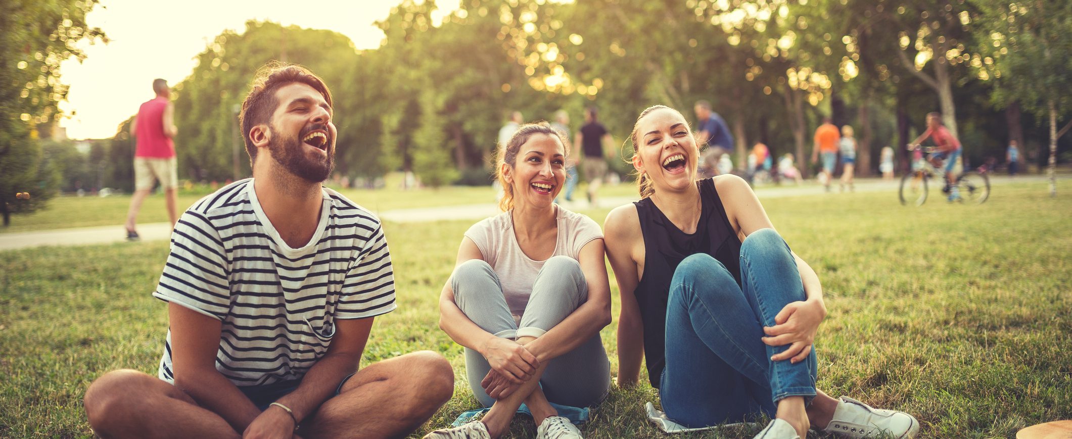 Three friends sitting on the grass in a park and laughing.
