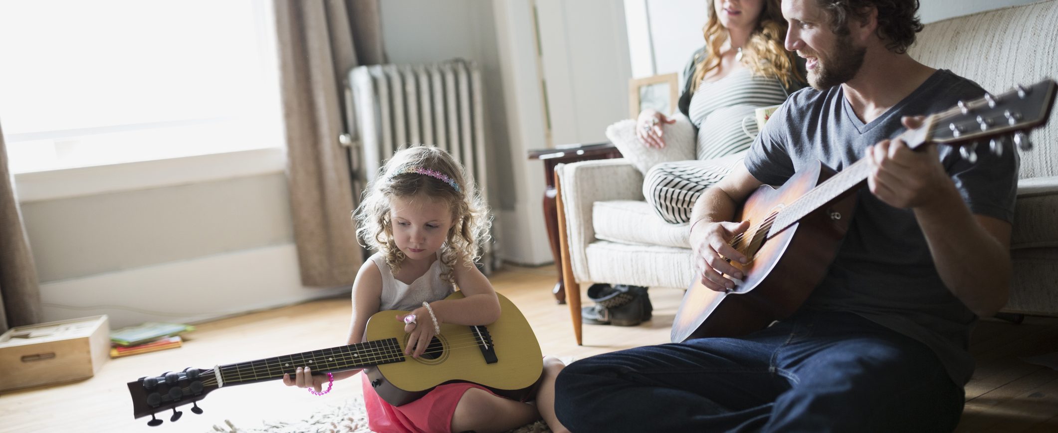 A man and a little girl playing guitars, while a woman sits on a couch.