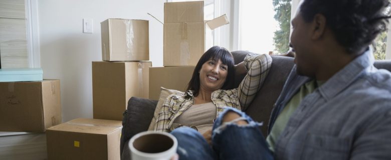 Two women relaxing and sipping coffee in the living room of their new home, surrounded by boxes.
