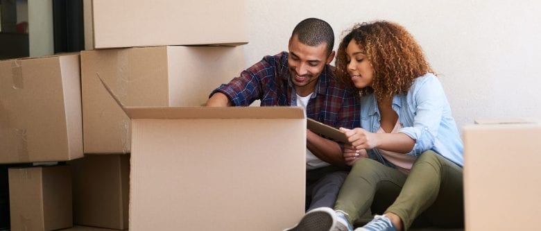 Two people read information off a clipboard as they sit among a lot of boxes