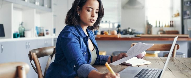 A woman, sitting at her kitchen table with a laptop computer, writes something in her notebook.