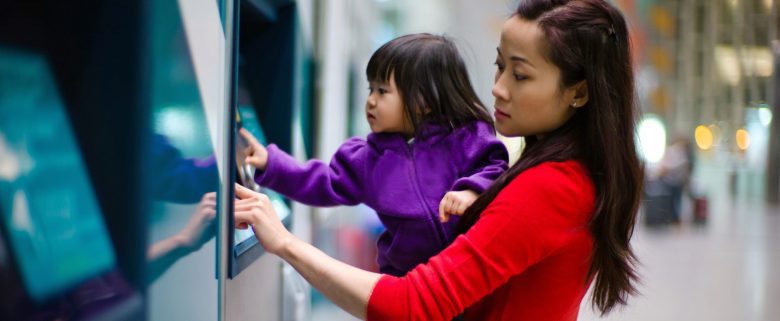 A woman holds a child while accessing an ATM machine. 