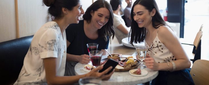 Three women seated at a restaurant table with food and drinks. One woman has her phone out and they’re all looking at it, laughing together.