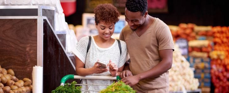 A couple looks at a smartphone together as they wheel a grocery cart through the produce section of a grocery store. 