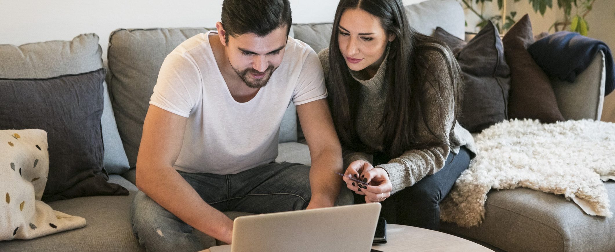 A man and woman sit on a couch together looking at laptop.