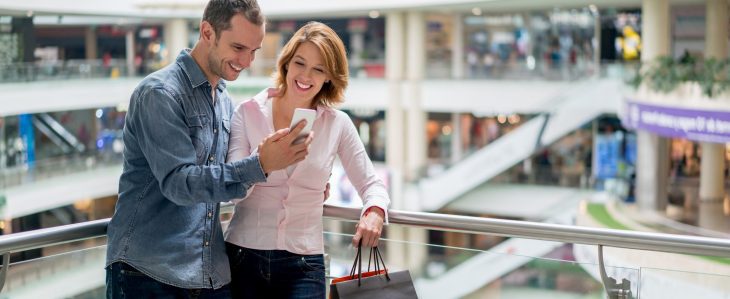 A man and woman lean against a railing at a mall looking at a phone together. 