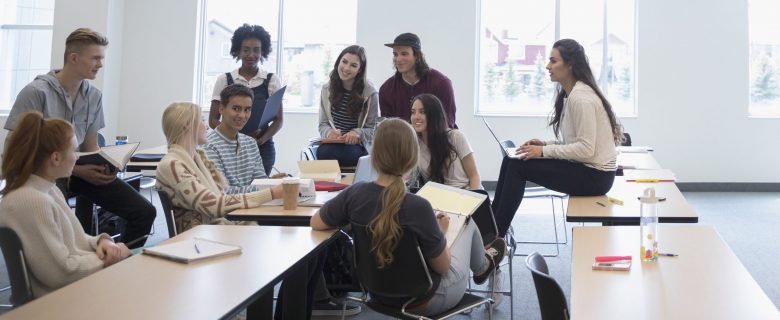 College students collaborate in a classroom together, sitting on desks and chairs with open laptops and notebooks.