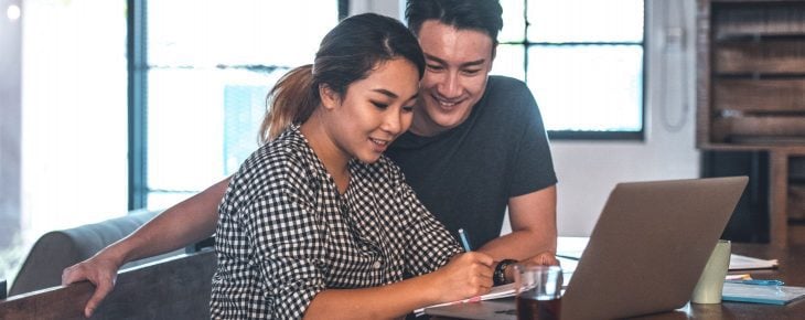 A couple looks at the screen of a laptop together while the woman also jots something in a notebook. 