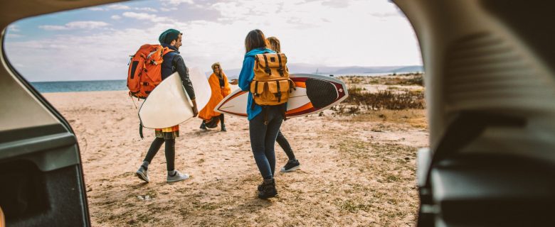 Four friends wearing backpacks gather on a beach holding surf boards.
