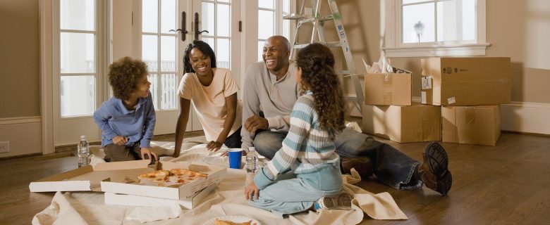 Family laughs together as they sit on the floor, eating pizza, in their new home.