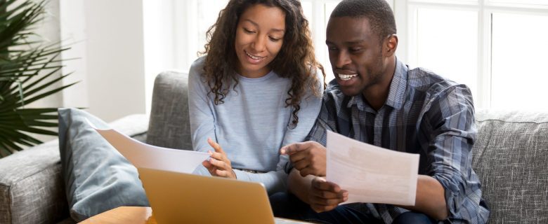 Two people on a sofa by the window sit together and talk about papers they’re holding while looking at a laptop.