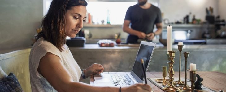 A woman sits at her kitchen table and works on her retirement budget, while her husband fixes dinner in the kitchen.