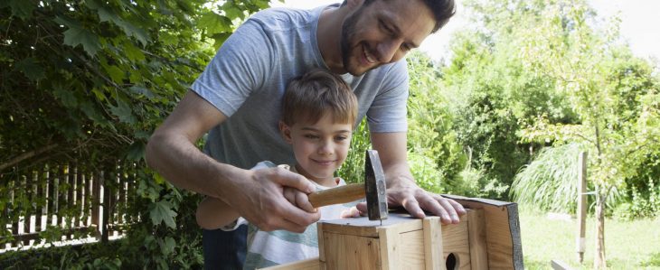 A dad and his son hammer a nail into a bird house. 