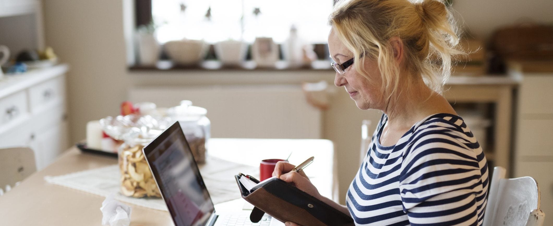 A woman writes in a notebook while looking at her laptop.