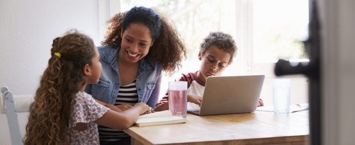 A mom sits at the kitchen table with her young son who is looking at a laptop and her daughter who is reading a book. 