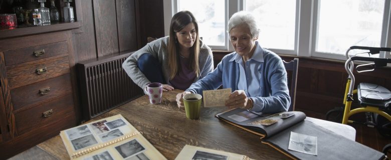 A person sits with an elderly woman at a wooden dining table with their coffee, looking over old scrapbooks and photos.