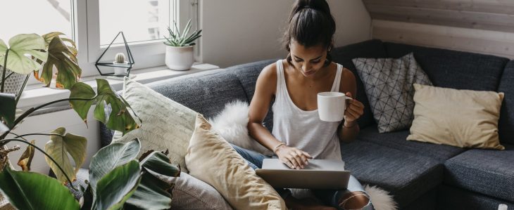 A woman drinking coffee on her couch types something into her laptop computer.
