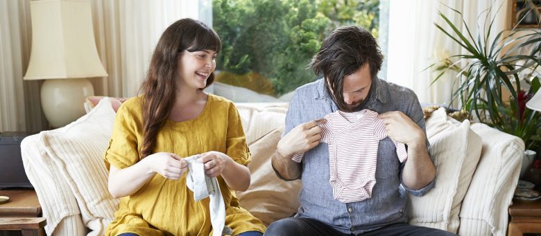 A man and a woman, sitting on their couch and looking at baby clothes.