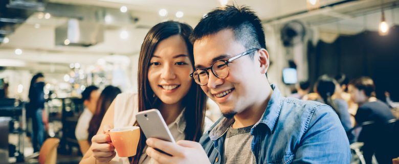 A smiling couple in a coffee shop looks at the screen of a smartphone. 