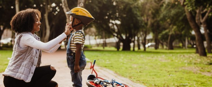 A smiling woman helps a little boy buckle his yellow helmet. A bicycle rests on its side beside them. 