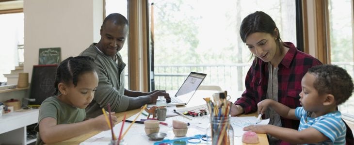 A family sits at a table together. The dad types on a laptop while the son and daughter draw as the mom smiles and looks at their work. 