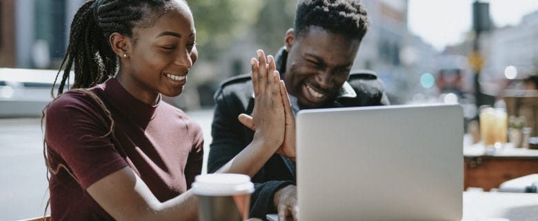 A man and a woman sit at an outdoor coffee shop, looking at a laptop and giving each other a high five. 