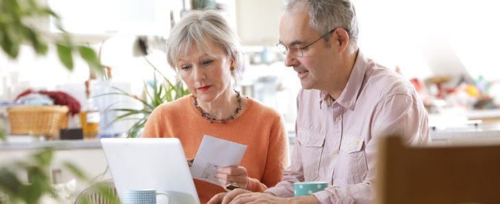 An older couple sit together at a kitchen table and look at a laptop screen together. 