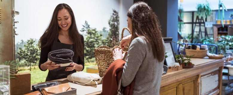 In a shop, one woman holds a blanket while another woman holds a basket.