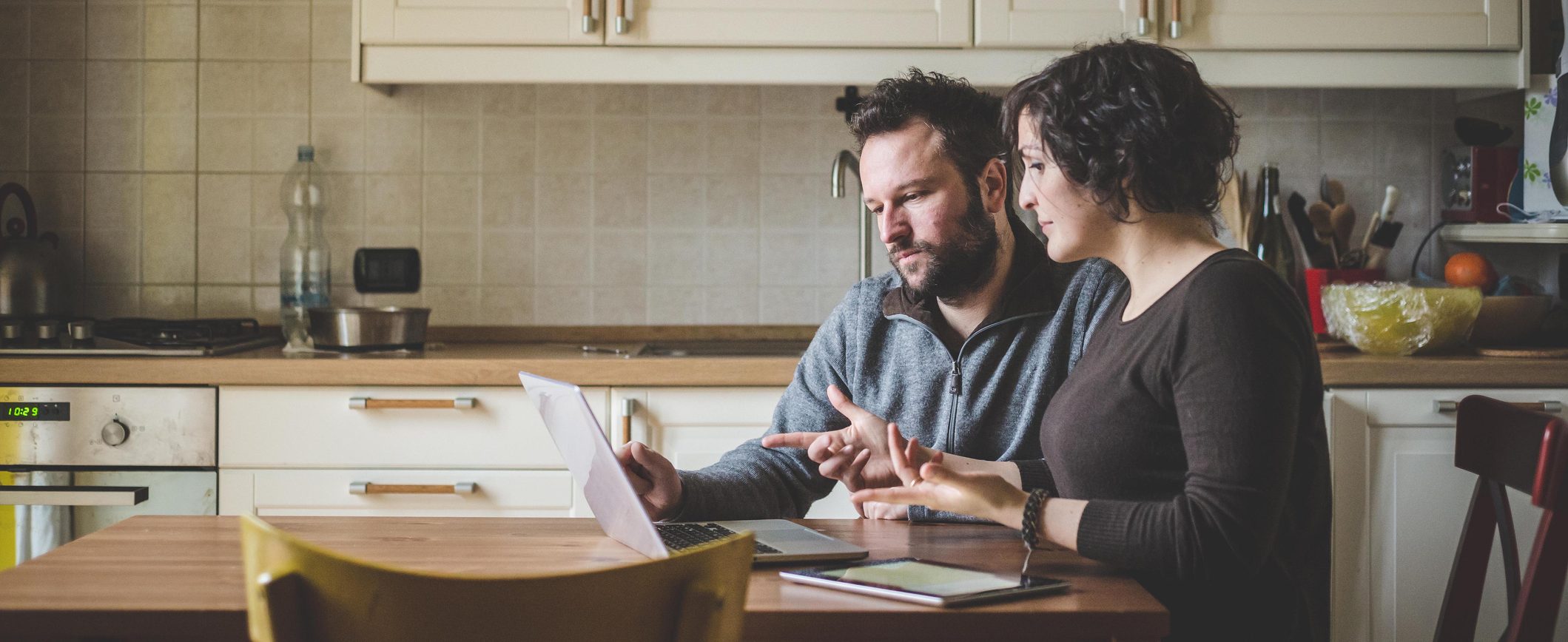 A married couple sitting at their kitchen table, discussing their emergency finance plan as they look at a lap computer screen.