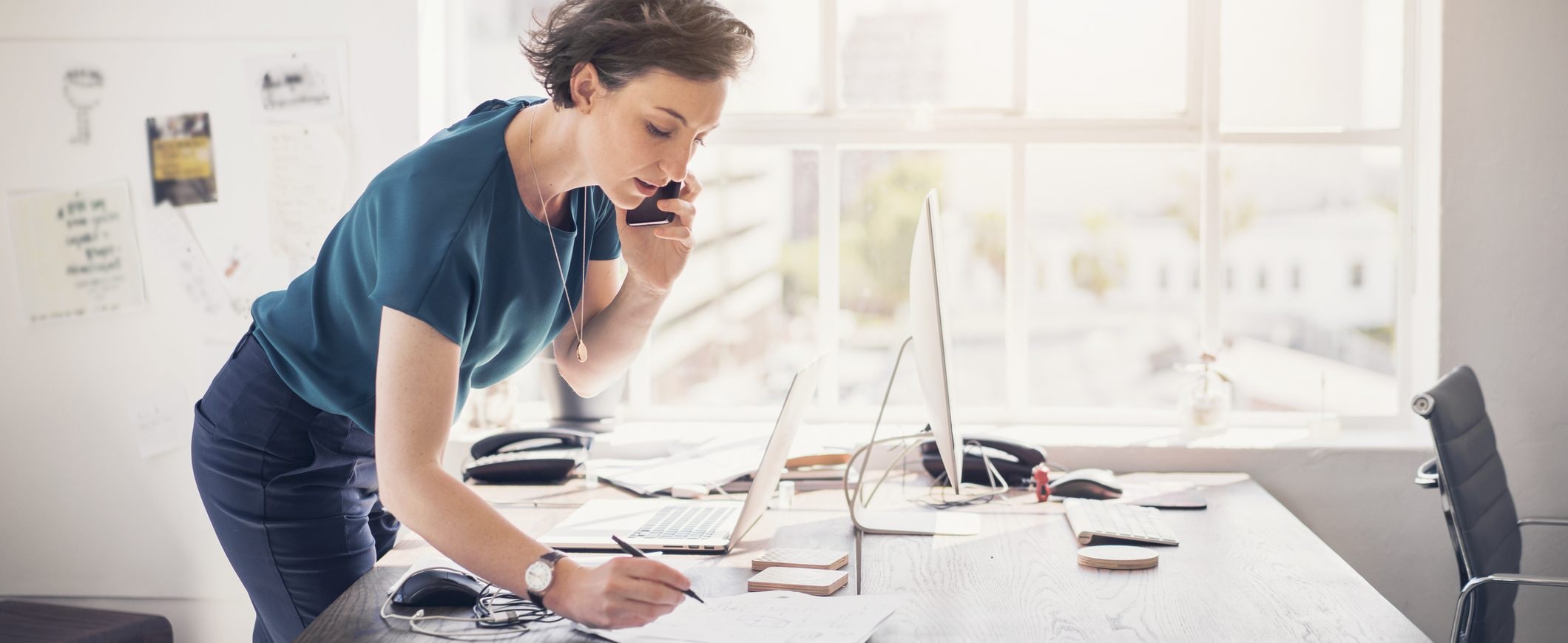 A woman on the phone, standing at her desk, taking notes with a pen and paper.