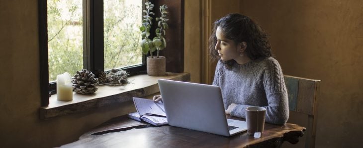 A woman sits at a table with an outside window, presumably studying as she sips a cup of tea.