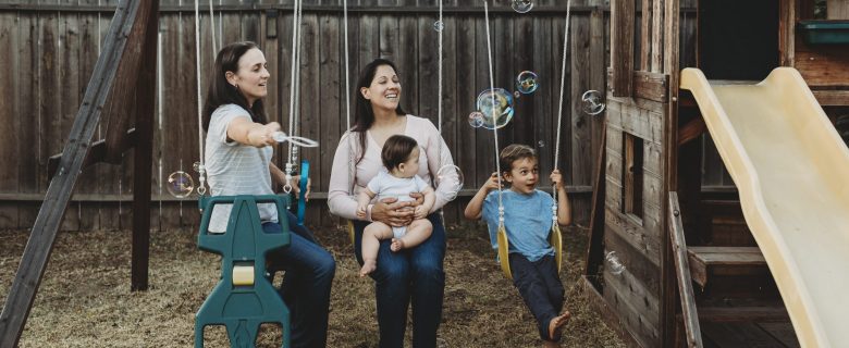 Two women, sitting on swings with their young children and blowing bubbles.
