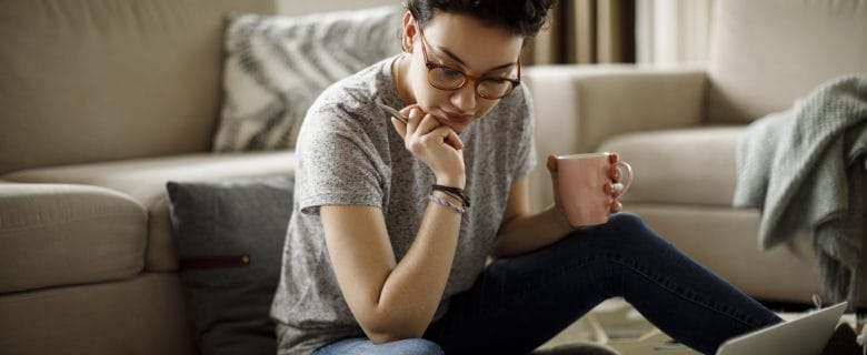 A woman sitting on her living room floor, drinking coffee and looking at her laptop computer.