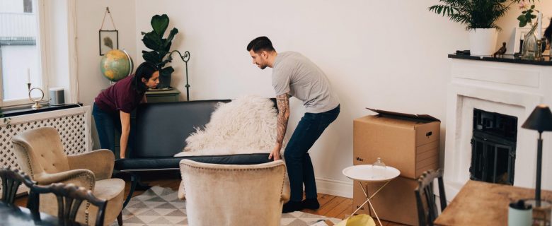 A man and a woman lift a couch together in a room. 