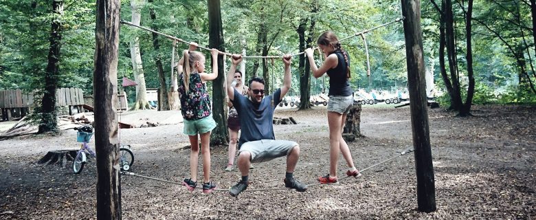 A family of four enjoys a day in a wooded park, climbing on a slack line.