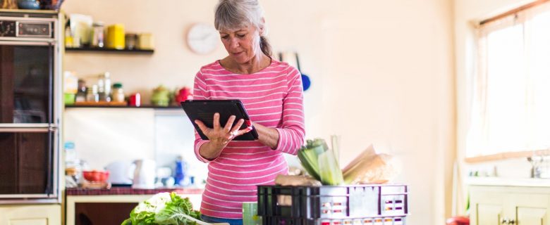 A woman looks at her tablet device while standing in the kitchen.