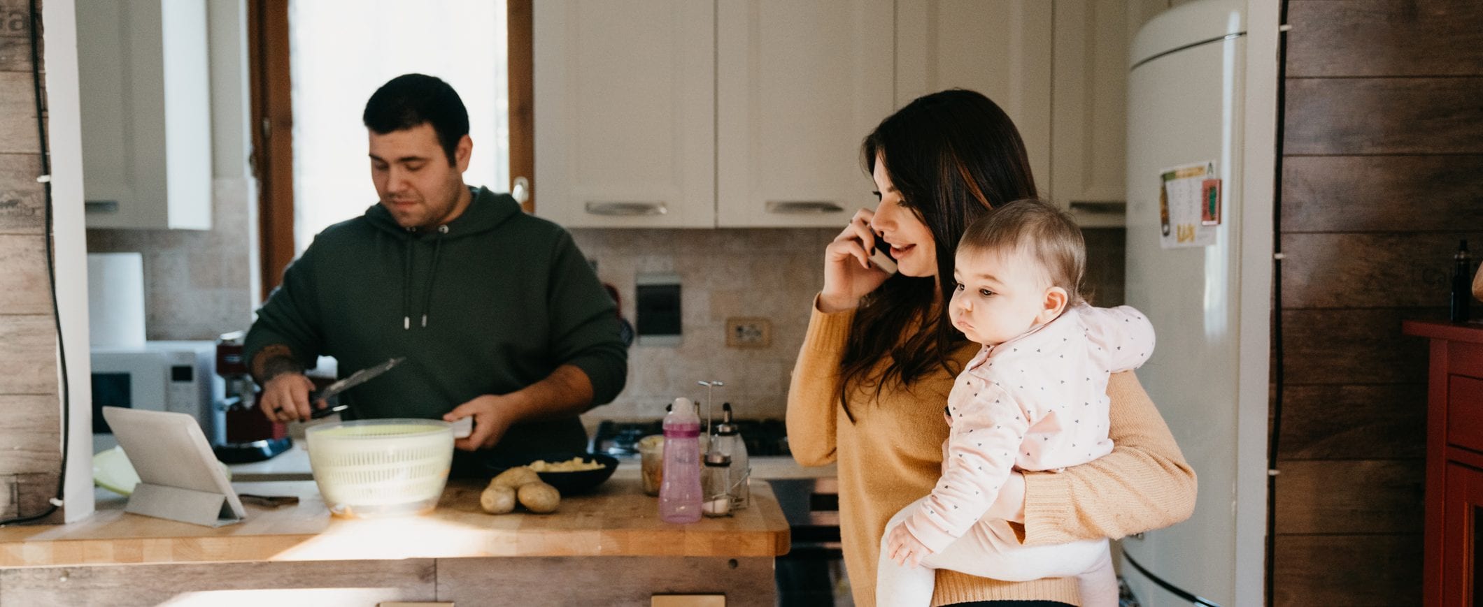 A man and woman stand in the kitchen. The man is preparing a meal and the woman is talking on the phone while holding her baby.