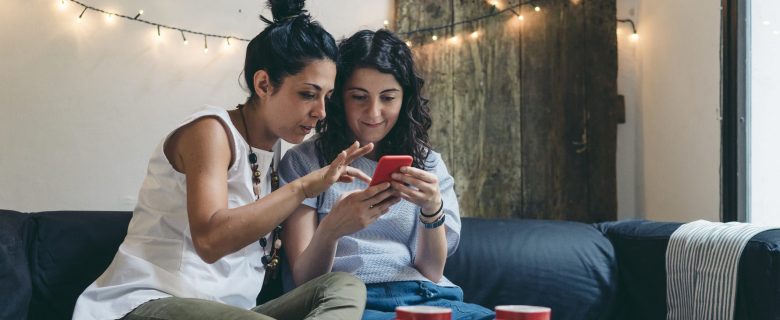 Two women sit on a couch looking at a mobile device.