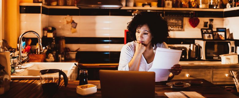 A woman sits in her kitchen looking at a laptop and holding a piece of paper.