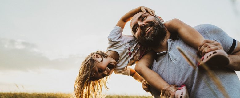 A little girl laughs as she’s carried around on the shoulders of a smiling man.