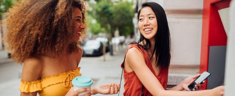 Two young women laughing together while one gets money from an ATM.